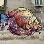 Un grand graffiti coloré sur un mur de béton représentant un poisson stylisé. Le corps du poisson est rempli de motifs de feuilles russes, de fleurs blanches et d'un paysage de plage avec des palmiers. De minuscules personnages illustrés semblent grimper et sauter tout autour de lui.
