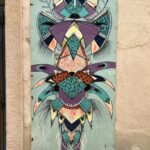 L'œuvre d'art datant de 2017 présente des motifs géométriques et organiques complexes ressemblant à un masque ou un totem abstrait, avec des formes symétriques et des couleurs vives comme le violet, le turquoise et le noir. La peinture est détaillée avec des hachures, des pois et des textures de bois visibles, et comprend également des éléments naturels tels que des formes de feuilles et de plumes, s'étendant verticalement.