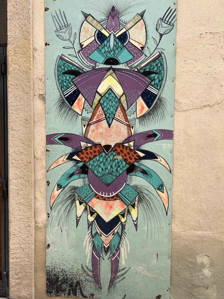 L'œuvre d'art datant de 2017 présente des motifs géométriques et organiques complexes ressemblant à un masque ou un totem abstrait, avec des formes symétriques et des couleurs vives comme le violet, le turquoise et le noir. La peinture est détaillée avec des hachures, des pois et des textures de bois visibles, et comprend également des éléments naturels tels que des formes de feuilles et de plumes, s'étendant verticalement.