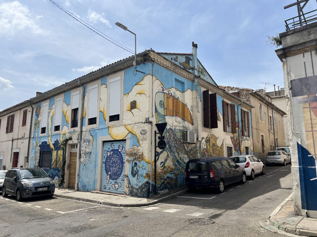 Photographie en contre-plongée d'une maison d'angle dans une rue en pente, entièrement recouverte d'une fresque de street art bleue et jaune représentant un toucan géant, des nuages et des vagues marines.