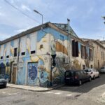 Photographie en contre-plongée d'une maison d'angle dans une rue en pente, entièrement recouverte d'une fresque de street art bleue et jaune représentant un toucan géant, des nuages et des vagues marines.